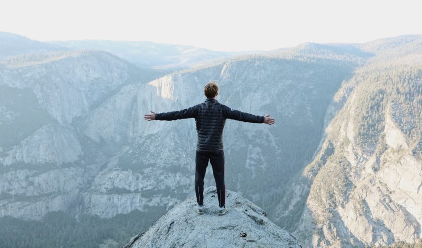 man opening his arms wide open on snow covered cliff with view of mountains during daytime